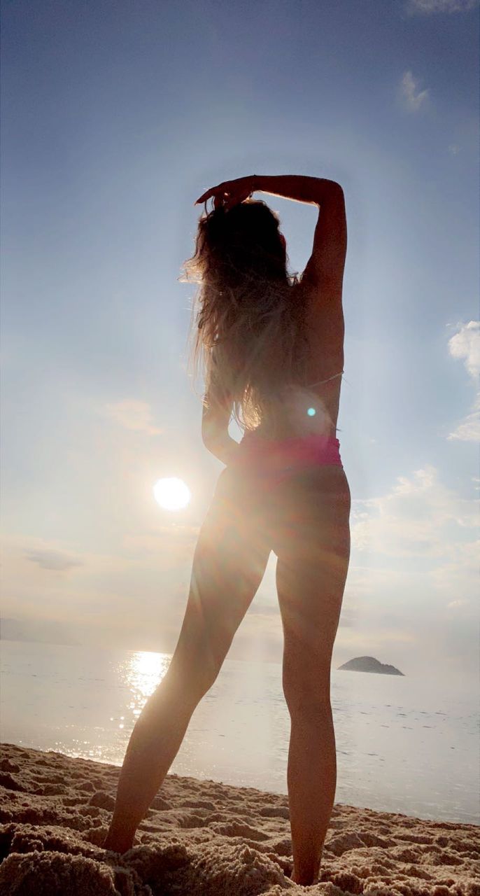 one person, adult, sky, women, land, nature, beach, sunlight, young adult, sea, water, full length, female, vacation, trip, hairstyle, clothing, rear view, holiday, standing, leisure activity, long hair, sunset, lifestyles, sand, back lit, beauty in nature, summer, lens flare, fashion, limb, outdoors, relaxation, sun, photo shoot, swimwear, cloud, brown hair, tranquility, human limb, environment, arm, arms raised, day, person, travel, carefree, copy space