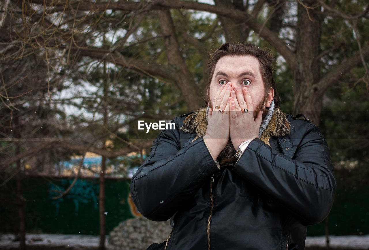 Shocked man with hands covering mouth against trees during winter at park