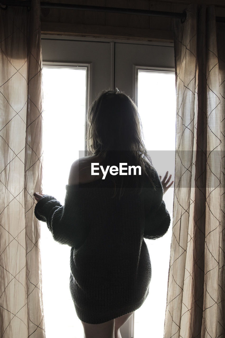 Rear view of woman standing amidst curtains in front of window