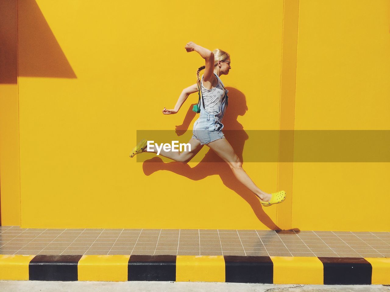Side view of mid adult woman jumping on footpath against yellow wall