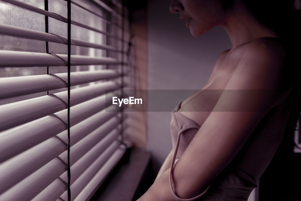 Midsection of sensuous woman wearing tank top while standing by window at home