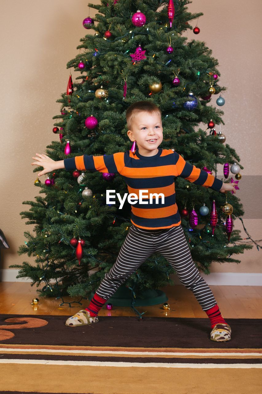 Smiling boy with arms outstretched standing against christmas tree at home