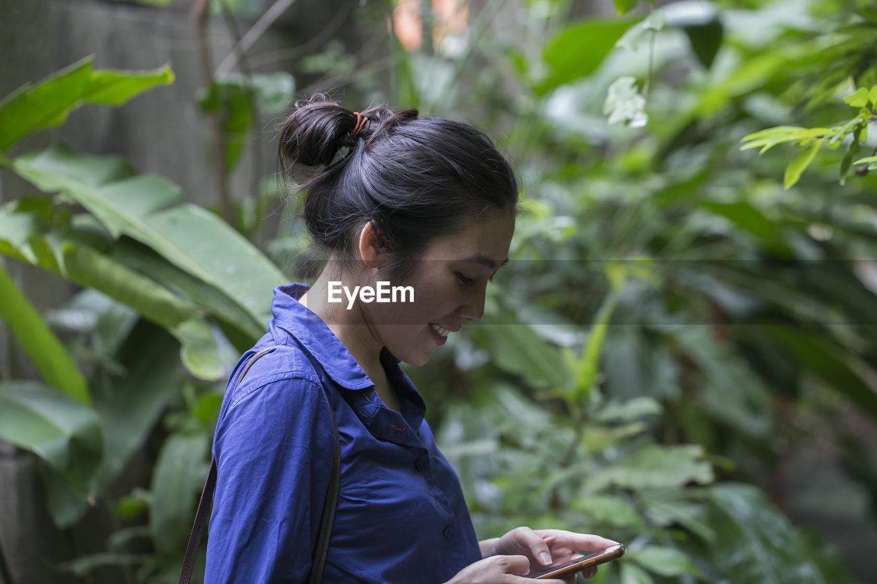 Side view of smiling young woman using mobile phone while standing against plants