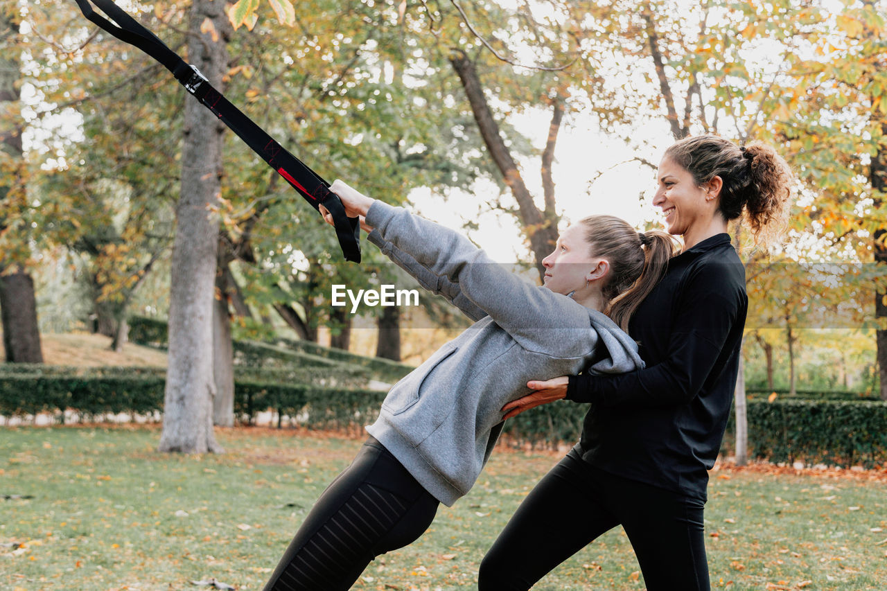 Happy friends exercising with straps while standing by tree