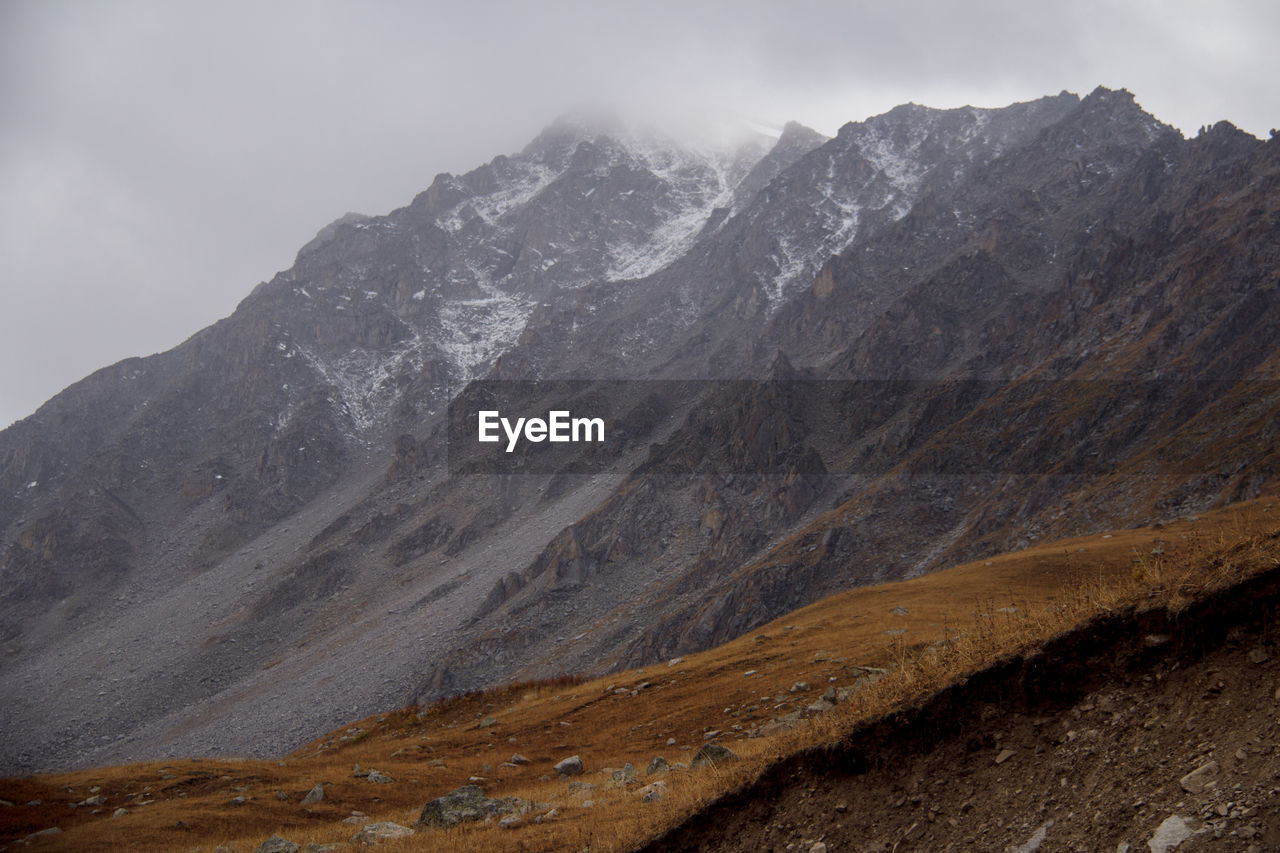 High rocky mountain with snow-capped peaks in fog against sky and clouds in bad weather in autumn