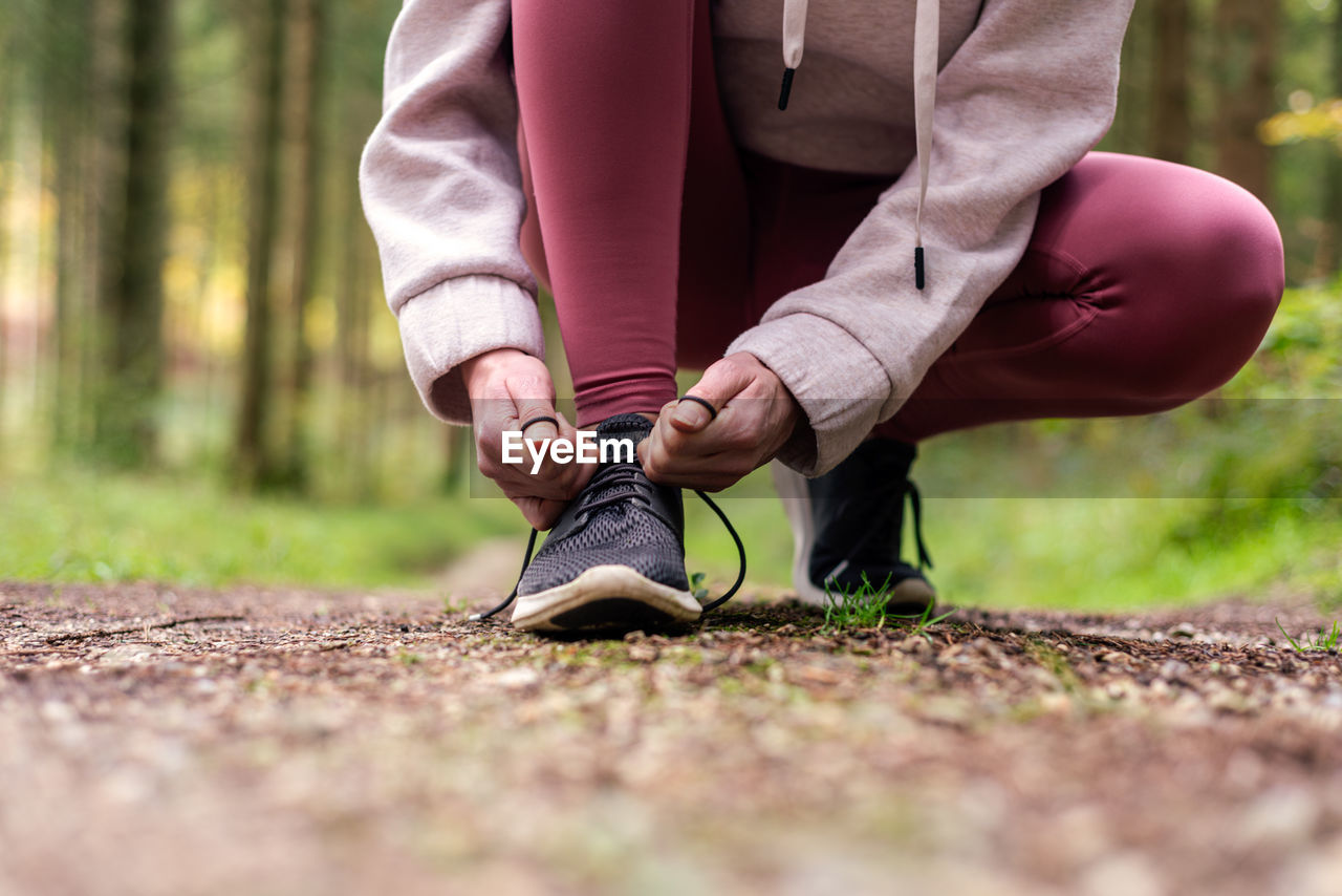 Cropped unrecognizable portswoman in activewear tying shoelaces on sneakers during workout in woods while having break on path