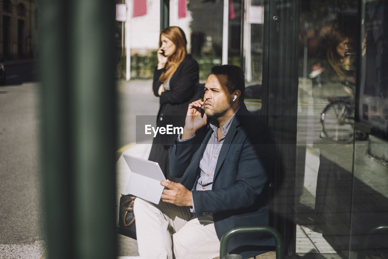 Male entrepreneur talking on smart phone while sitting with laptop at bus stop during sunny day