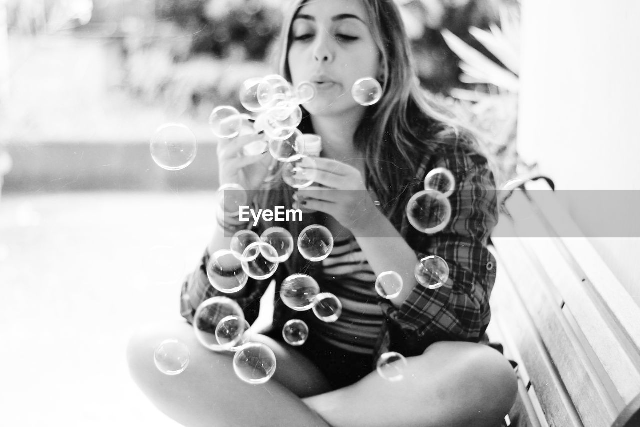 Young woman blowing soap bubbles while sitting on bench