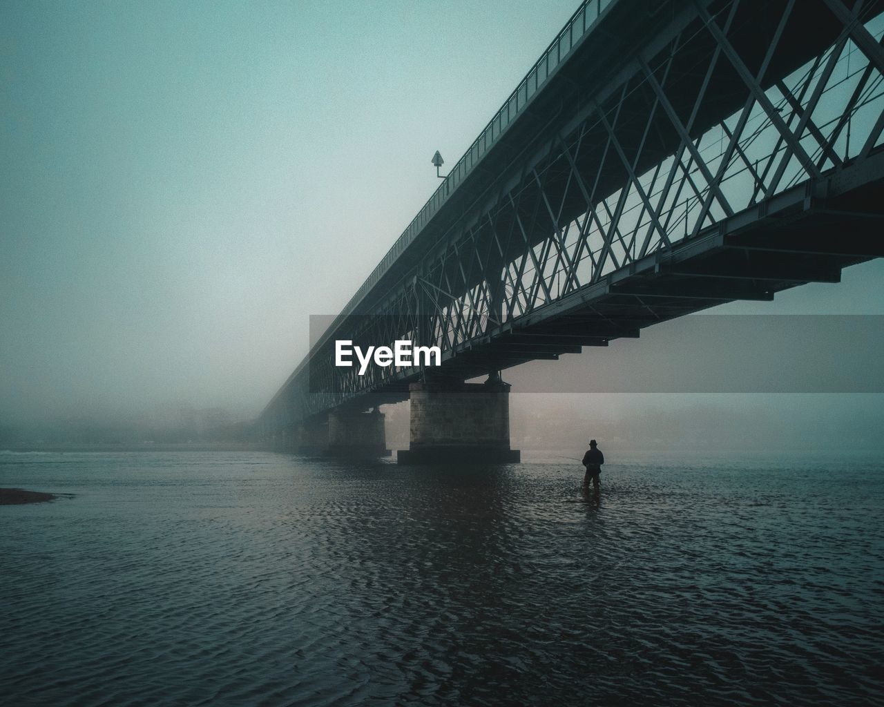 Silhouette person standing below bridge in lake against clear sky