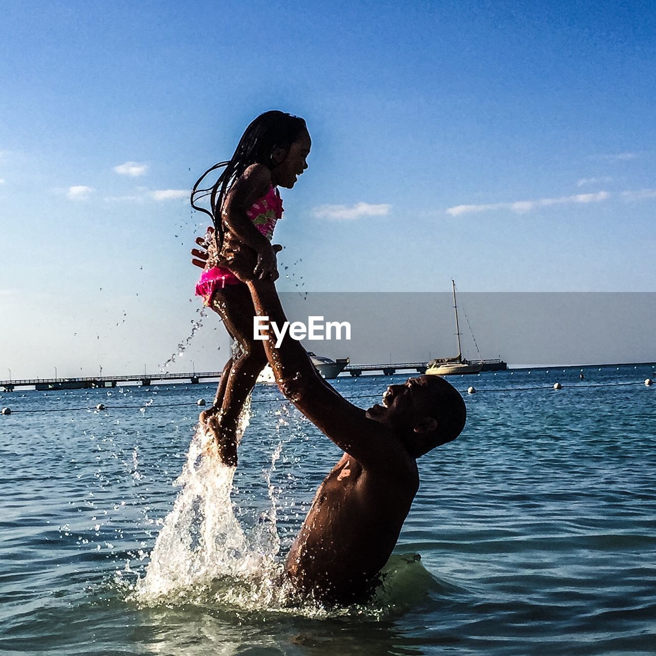 Side view of smiling father lifting daughter while standing in sea against sky