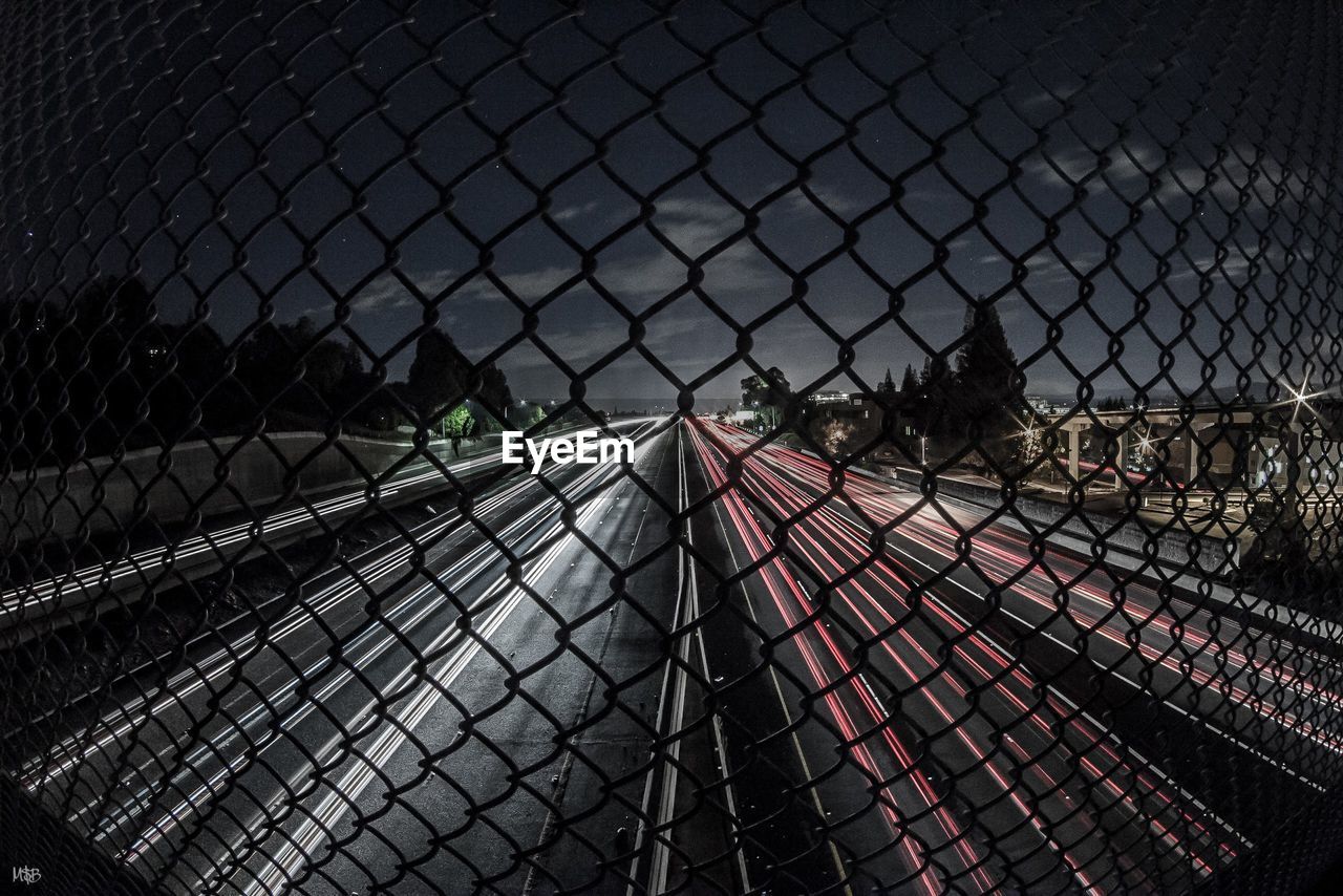 Light trails on road seen through chainlink fence at night