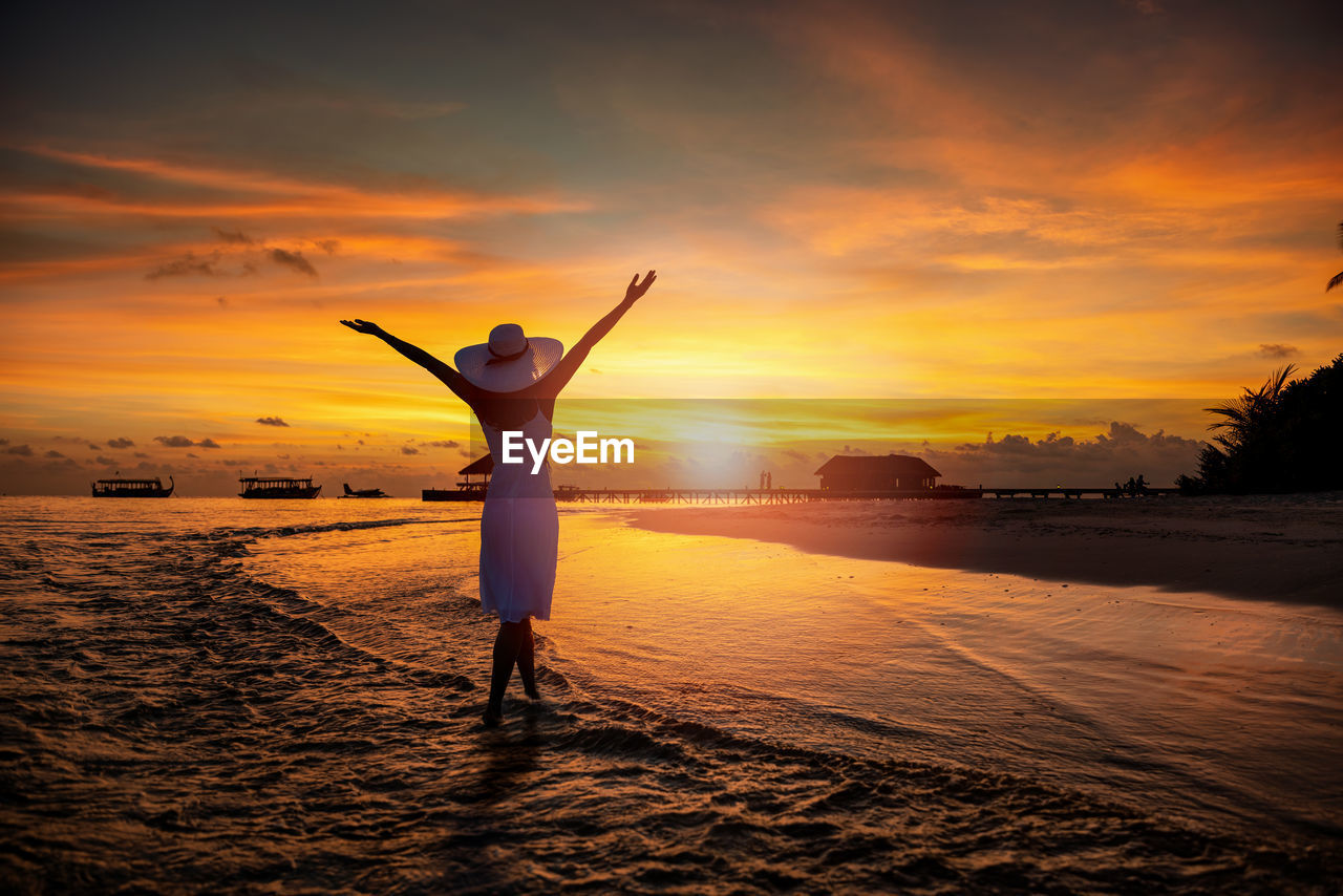 Rear view of woman with arms raised standing on shore at beach during sunset