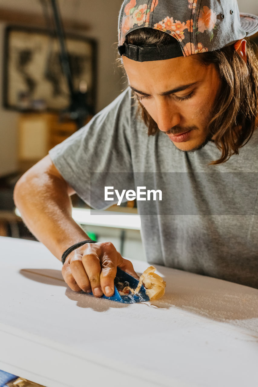 Focused male master using hand plane and shaping smooth surface of surfboard in workshop