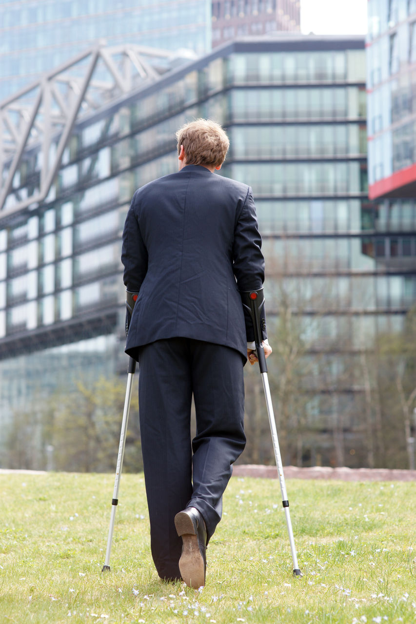 Businessman walks through a park in the city with crutches after knee injury