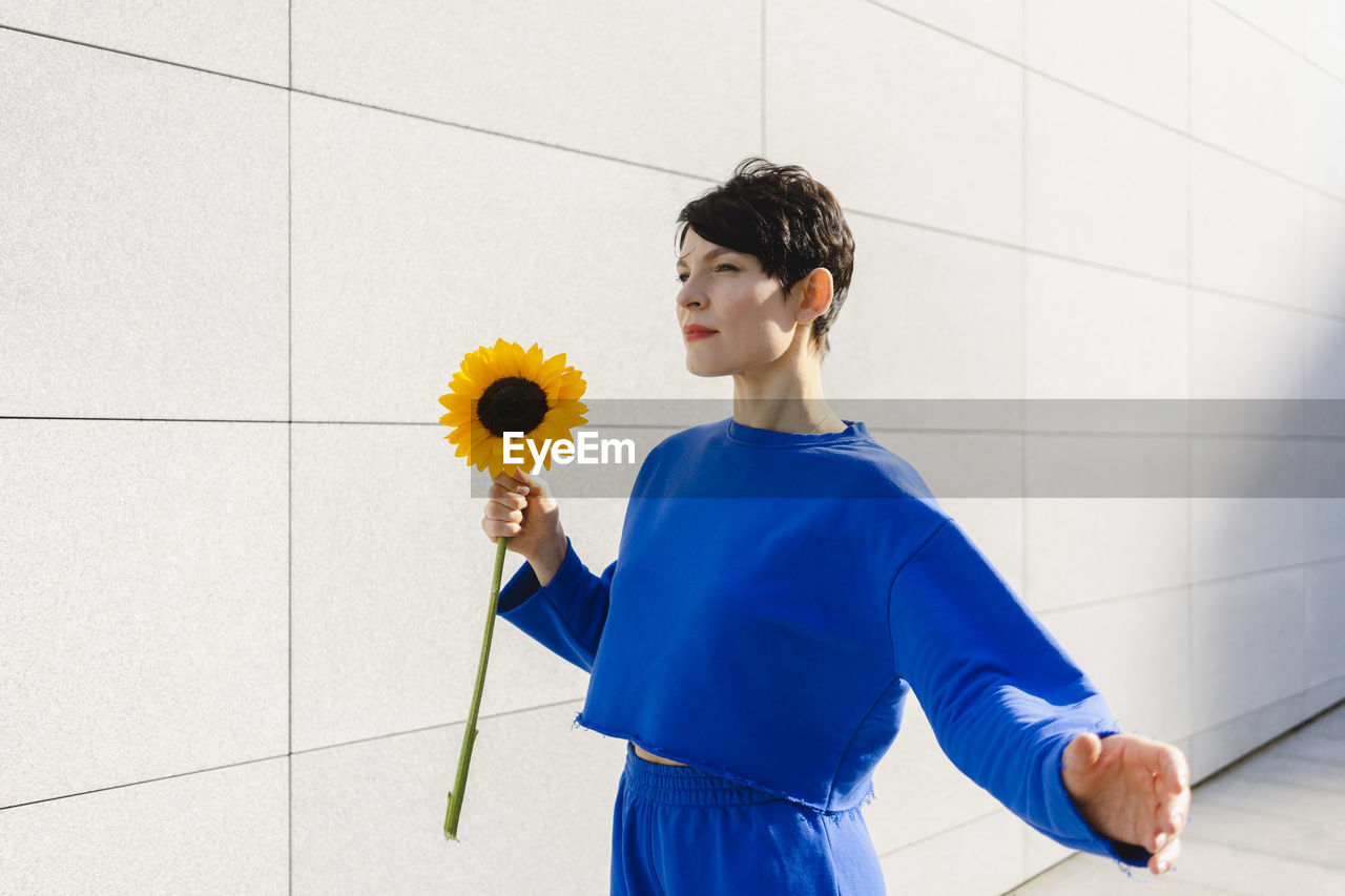 Woman with short hair holding sunflower by wall on sunny day