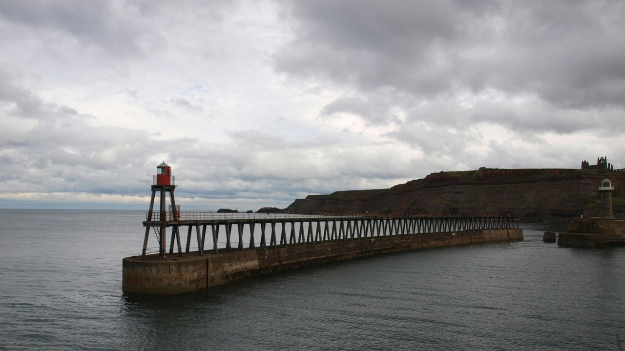 Beacon on pier in sea against cloudy sky