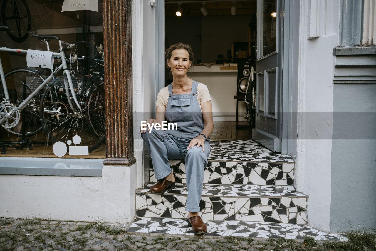 Portrait of confident senior owner sitting on steps of bicycle shop