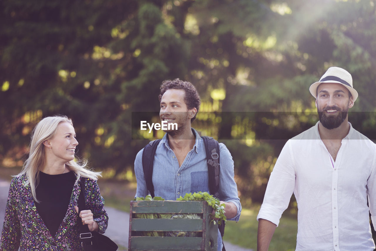 Smiling man holding crate while walking with friends against trees