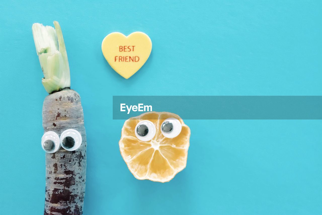 Directly above shot of heart shape with doodle eyes lemon and carrot against blue background