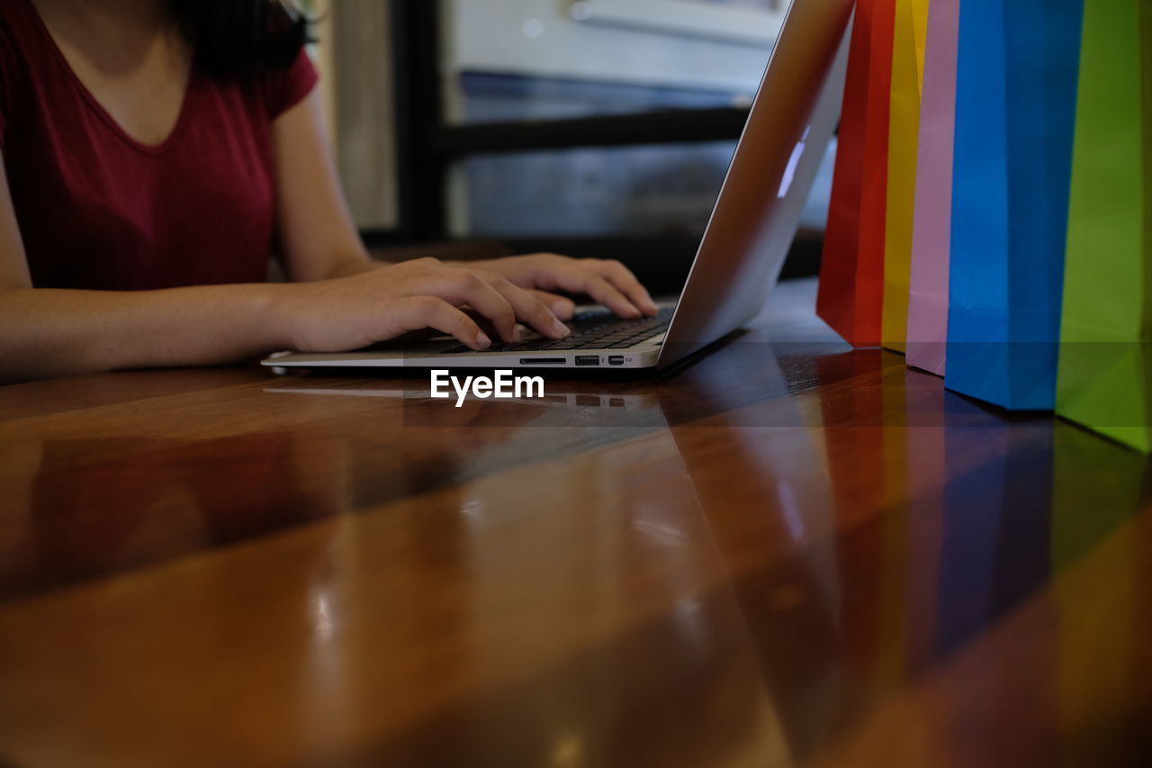 Close-up of woman hands using laptop on wooden table by colorful shopping bags