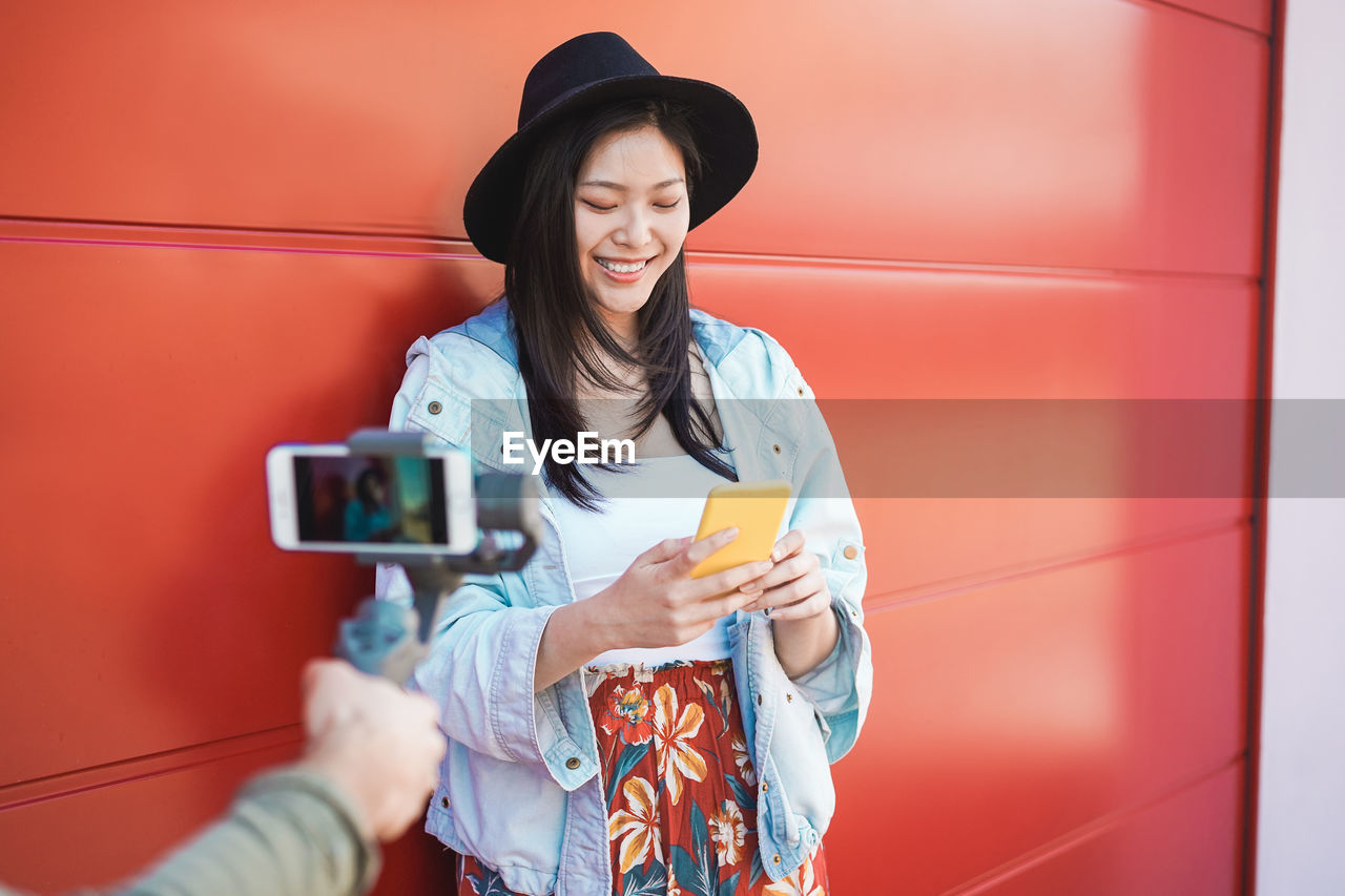 Happy young woman using mobile phone by cropped hand photographing against wall