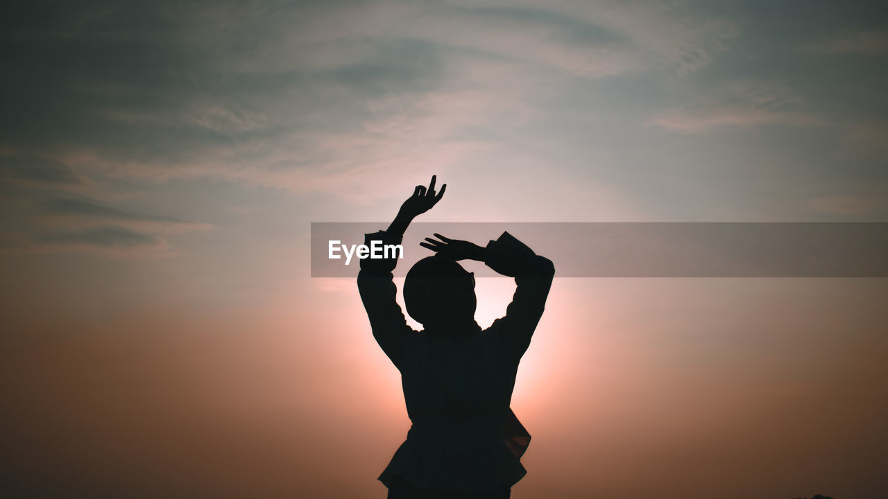 Silhouette woman with arms raised standing against sky during sunset