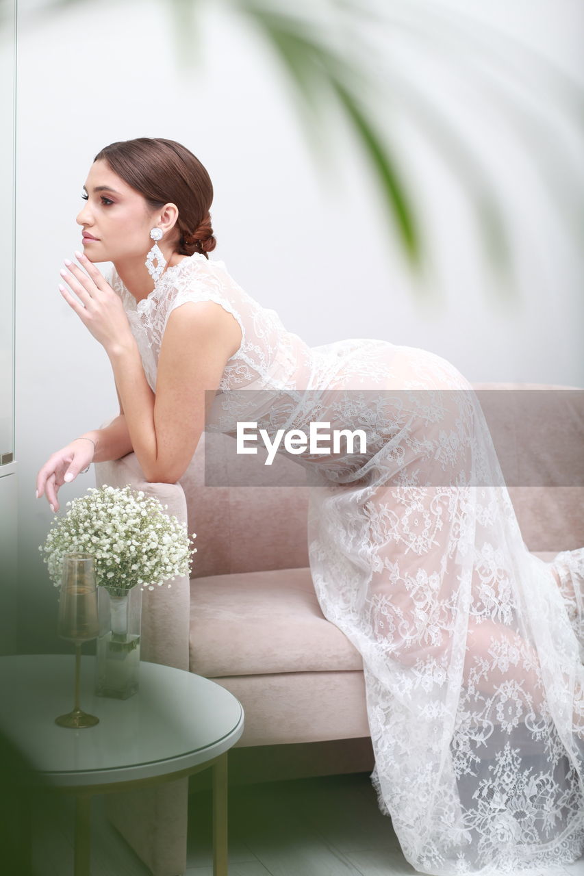 Side view portrait of bride in transparent lace night gown bending in sexy pose over armchair indoor