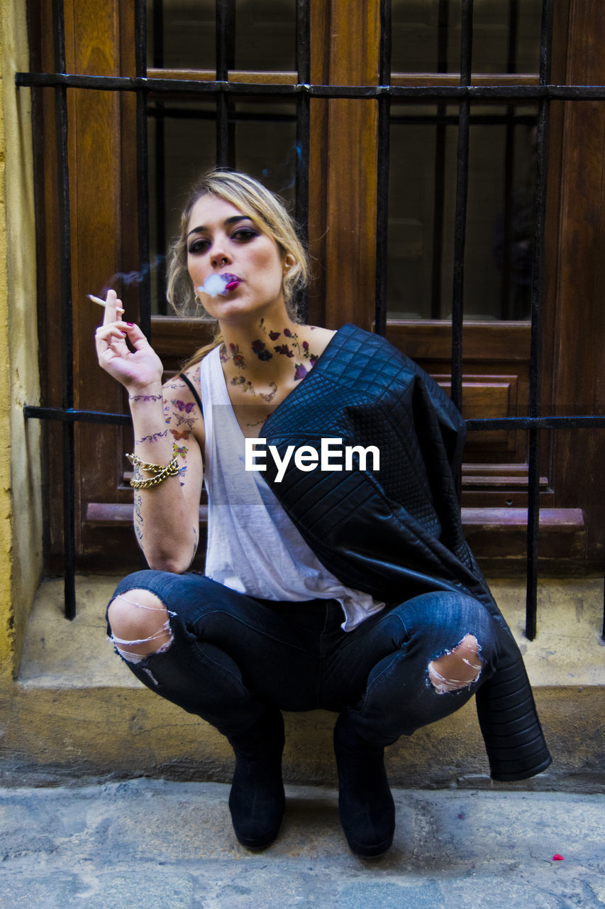 Portrait of young woman smoking cigarette while sitting in city