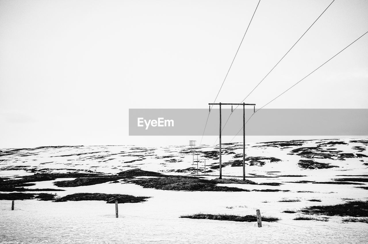 Electricity pylons over snow covered field against clear sky