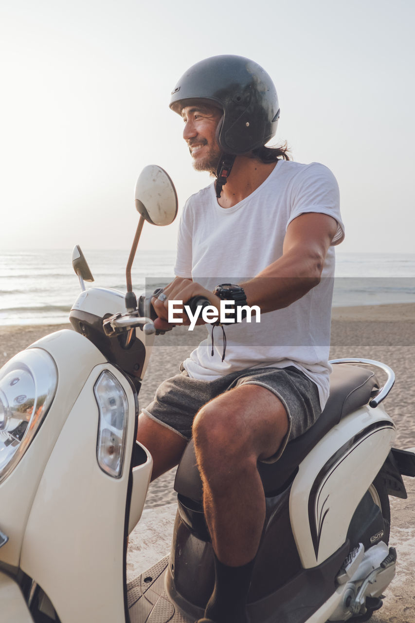 Side view of happy active guy dressed in white t shirt with shorts and black helmet riding scooter in summer evening on beach