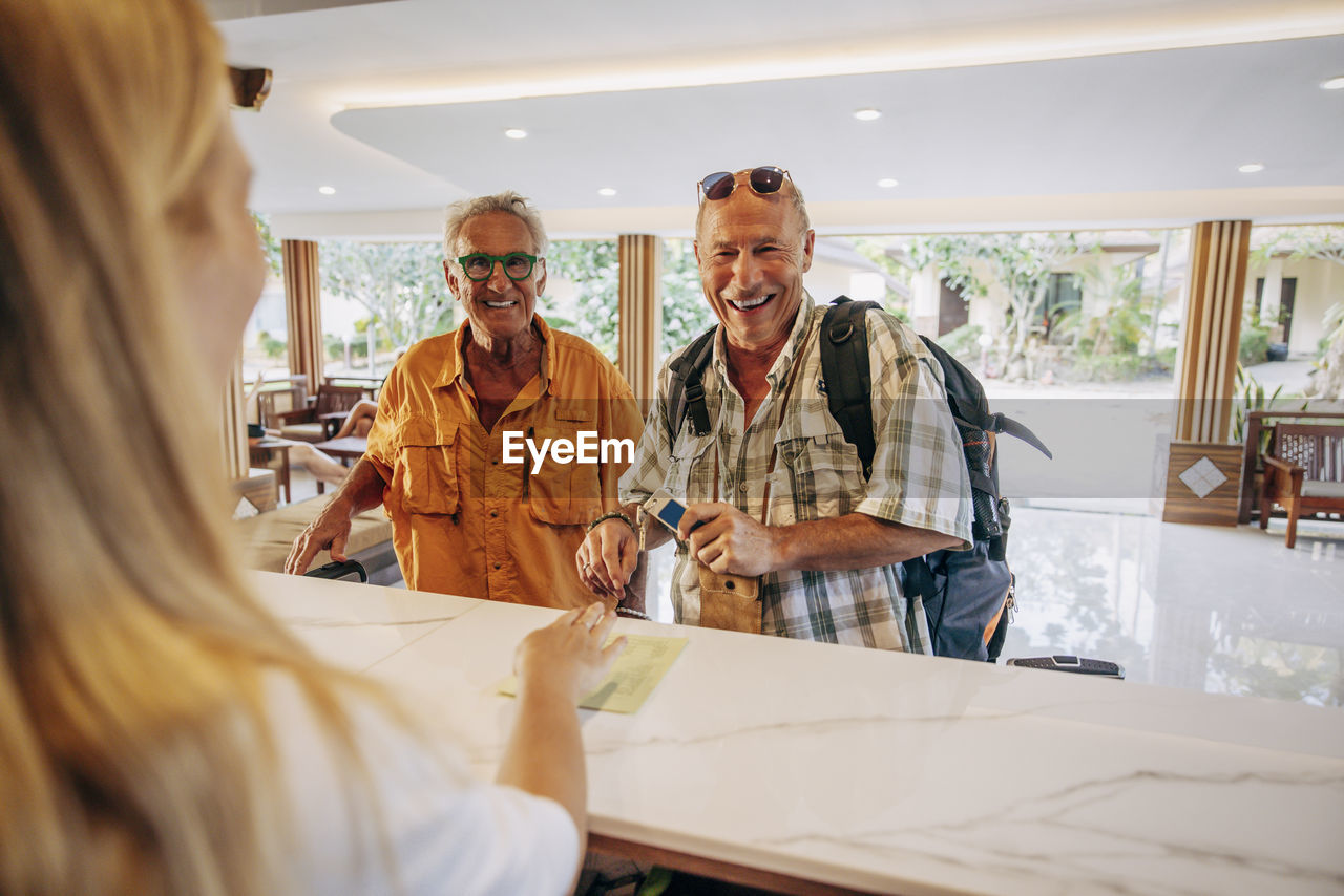 Happy senior gay couple talking with female receptionist at desk in hotel