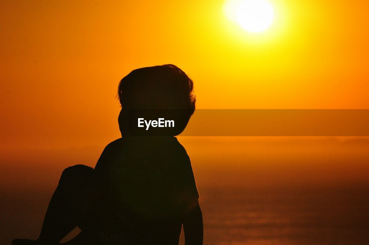 Silhouette boy sitting against sea during sunset