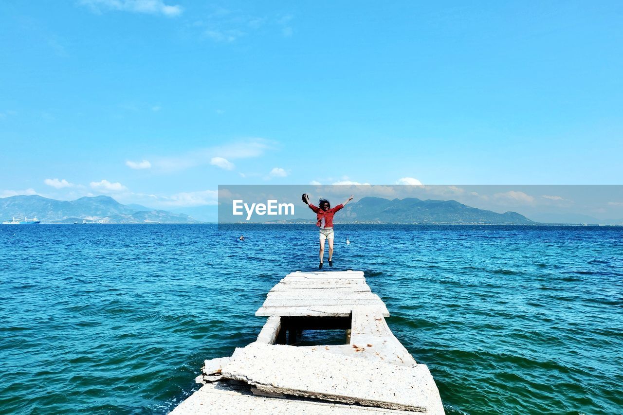 Woman jumping on old pier on sea against blue sky