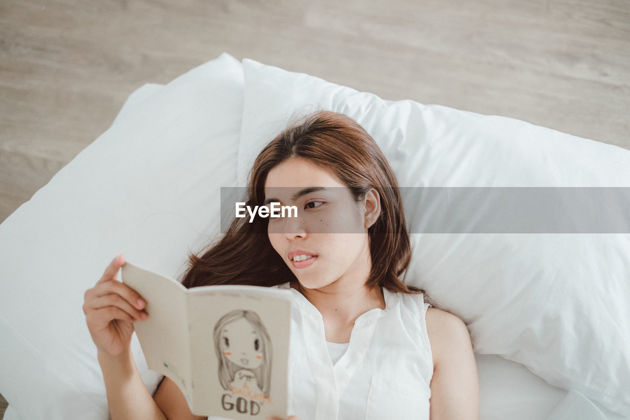 Smiling young woman reading book while lying on bed