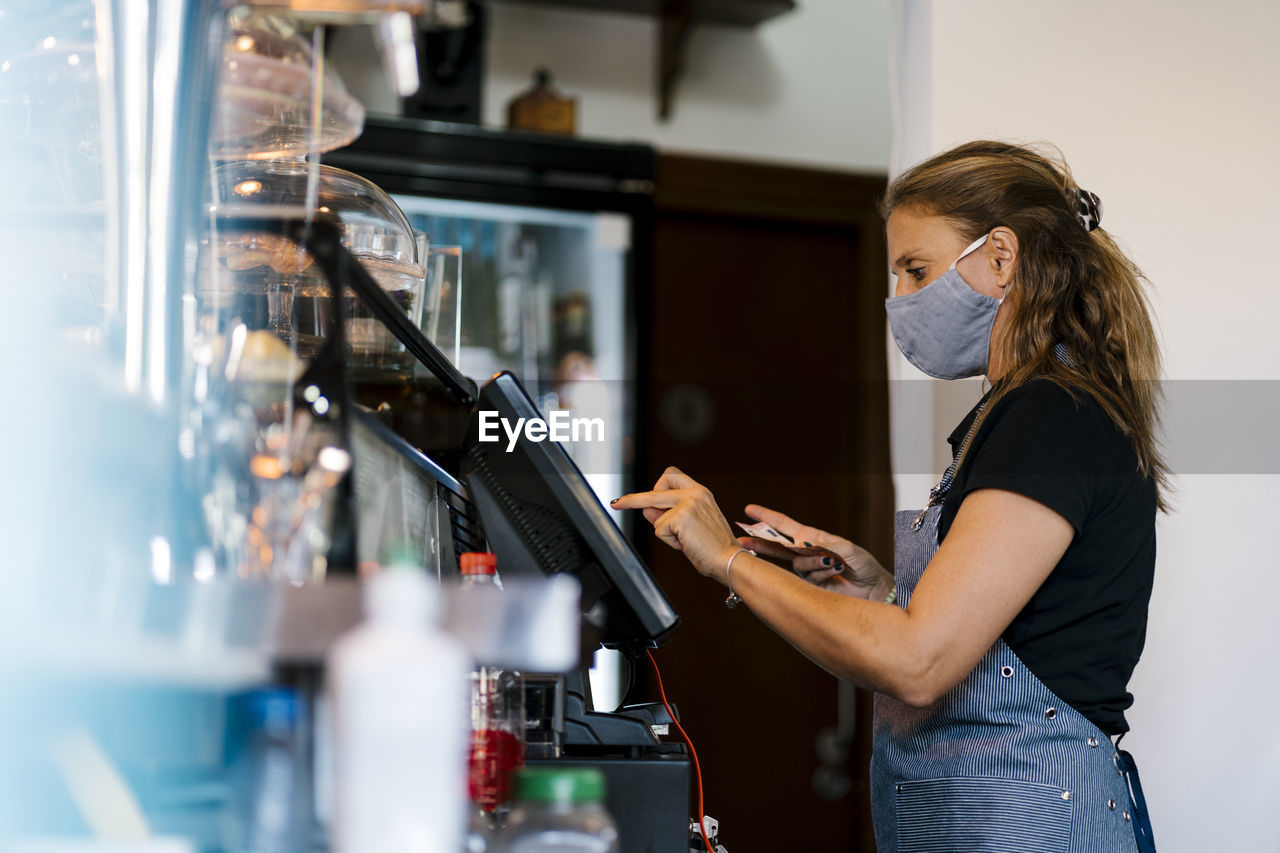 Female owner in protective face mask using cash register at coffee shop