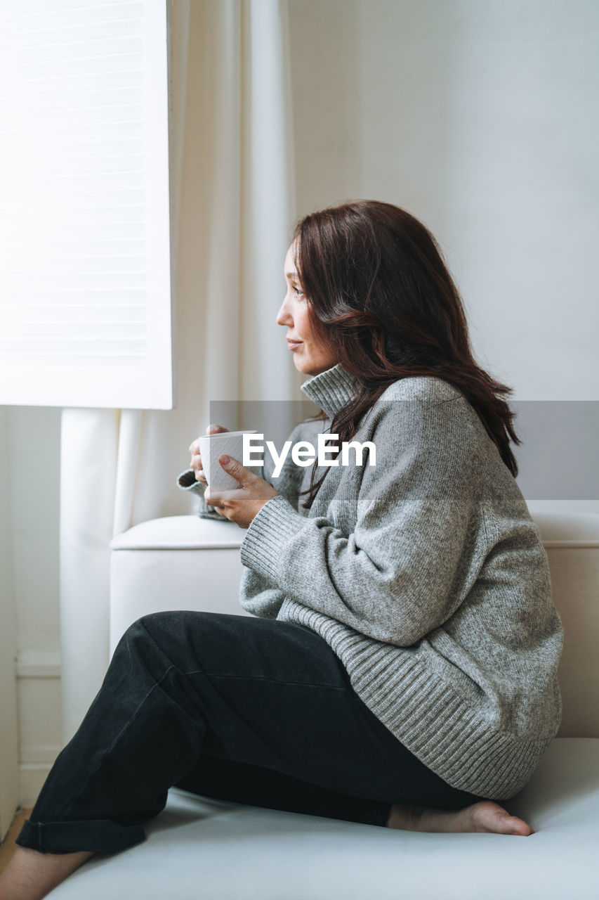 Thinking woman with brunette long hair in cozy knitted sweater with cup of tea in hands at home