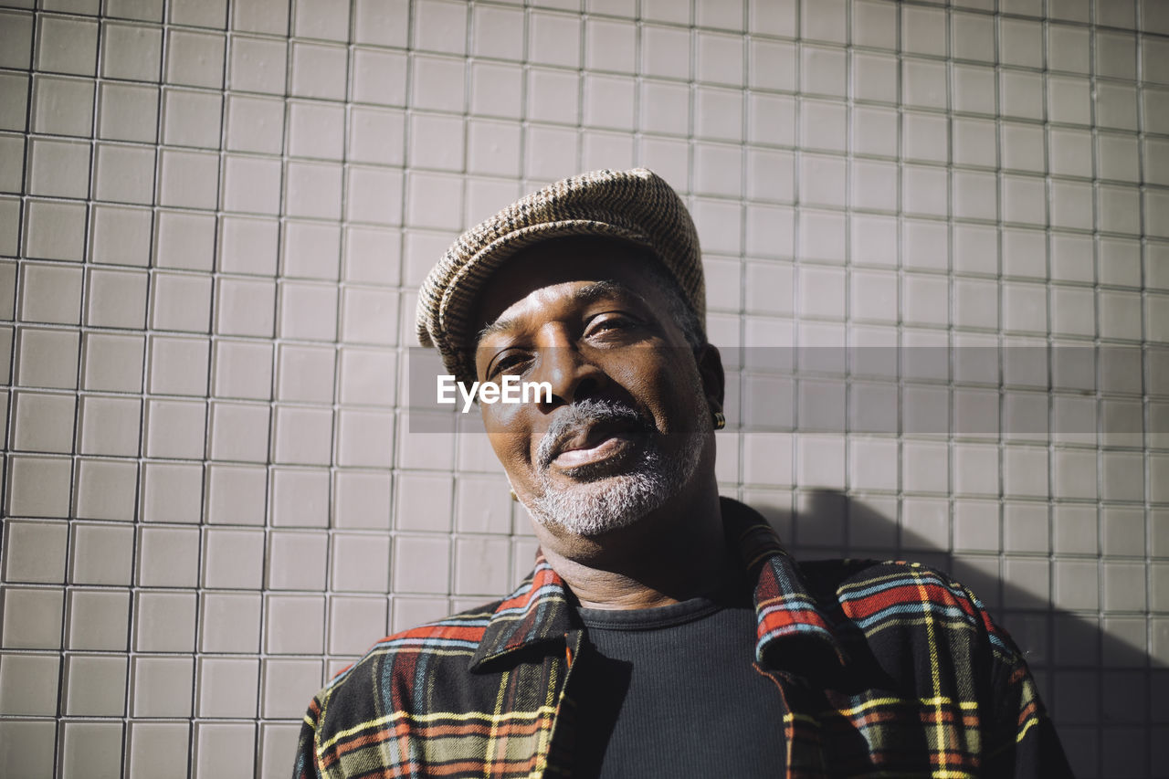 Portrait of smiling mature man wearing flat cap standing against wall