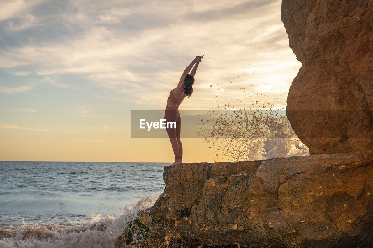 Woman standing on rocks by the sea doing a backbend, yoga pose.