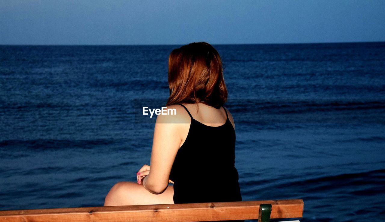 Woman sitting on bench at beach against clear sky