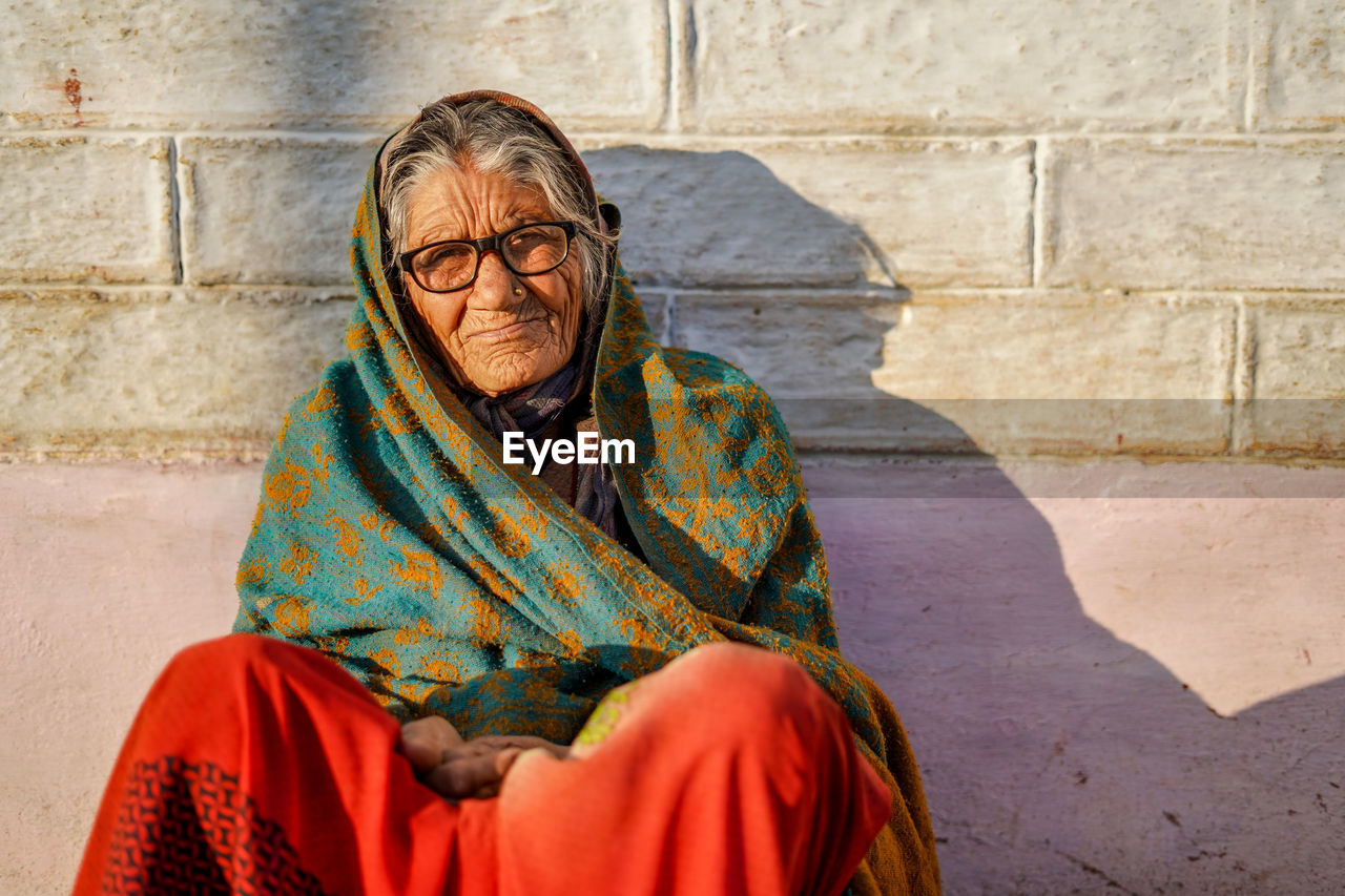 Old aged woman sitting with the support of the wall in the sunset wearing glasses