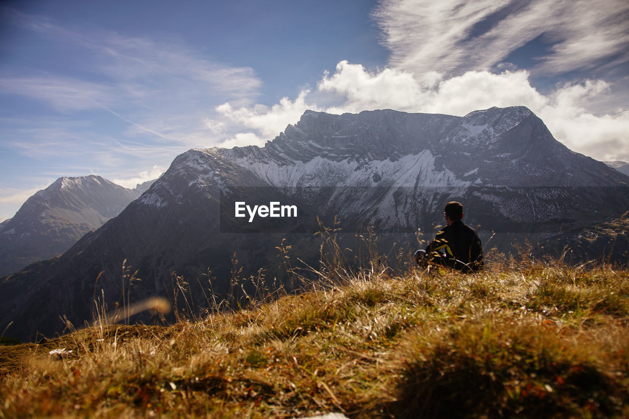 Man sitting on grassy field against mountains during sunny day