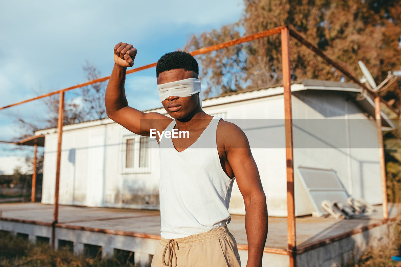 Anonymous masculine ethnic male protester in undershirt and blindfold standing with raised arm in countryside