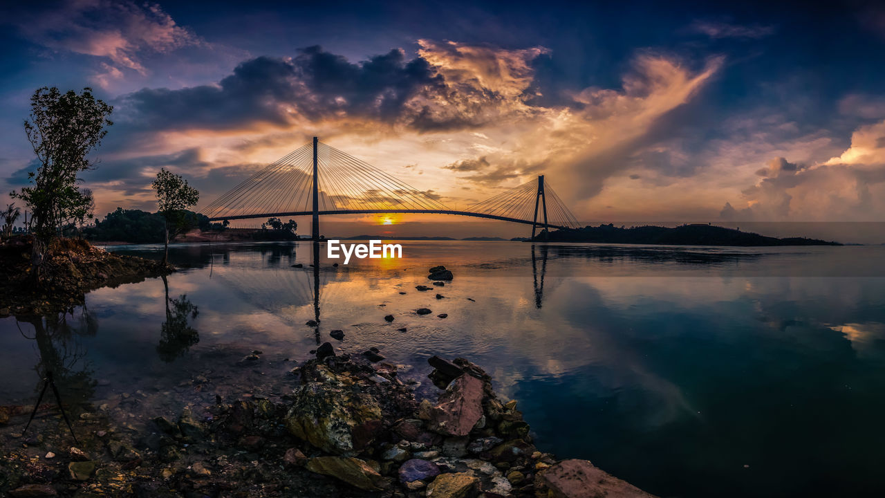 Silhouette barelang bridge over river against cloudy sky during sunset