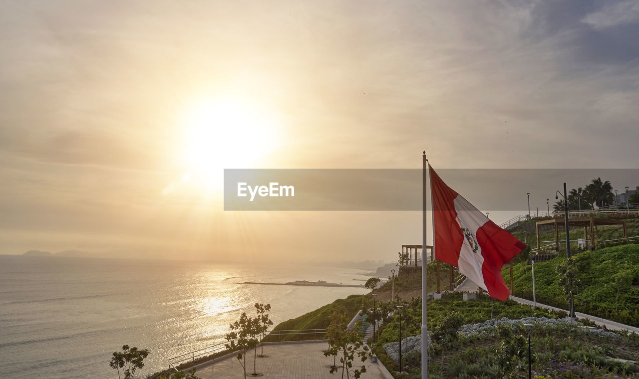 sky, flag, water, nature, sunset, patriotism, sea, sunlight, sun, beach, land, environment, cloud, beauty in nature, scenics - nature, tranquility, lens flare, architecture, travel destinations, outdoors, tranquil scene, coast, no people, ocean, travel, sunbeam, built structure, horizon over water, red, horizon, day, idyllic, wind, tourism, transportation