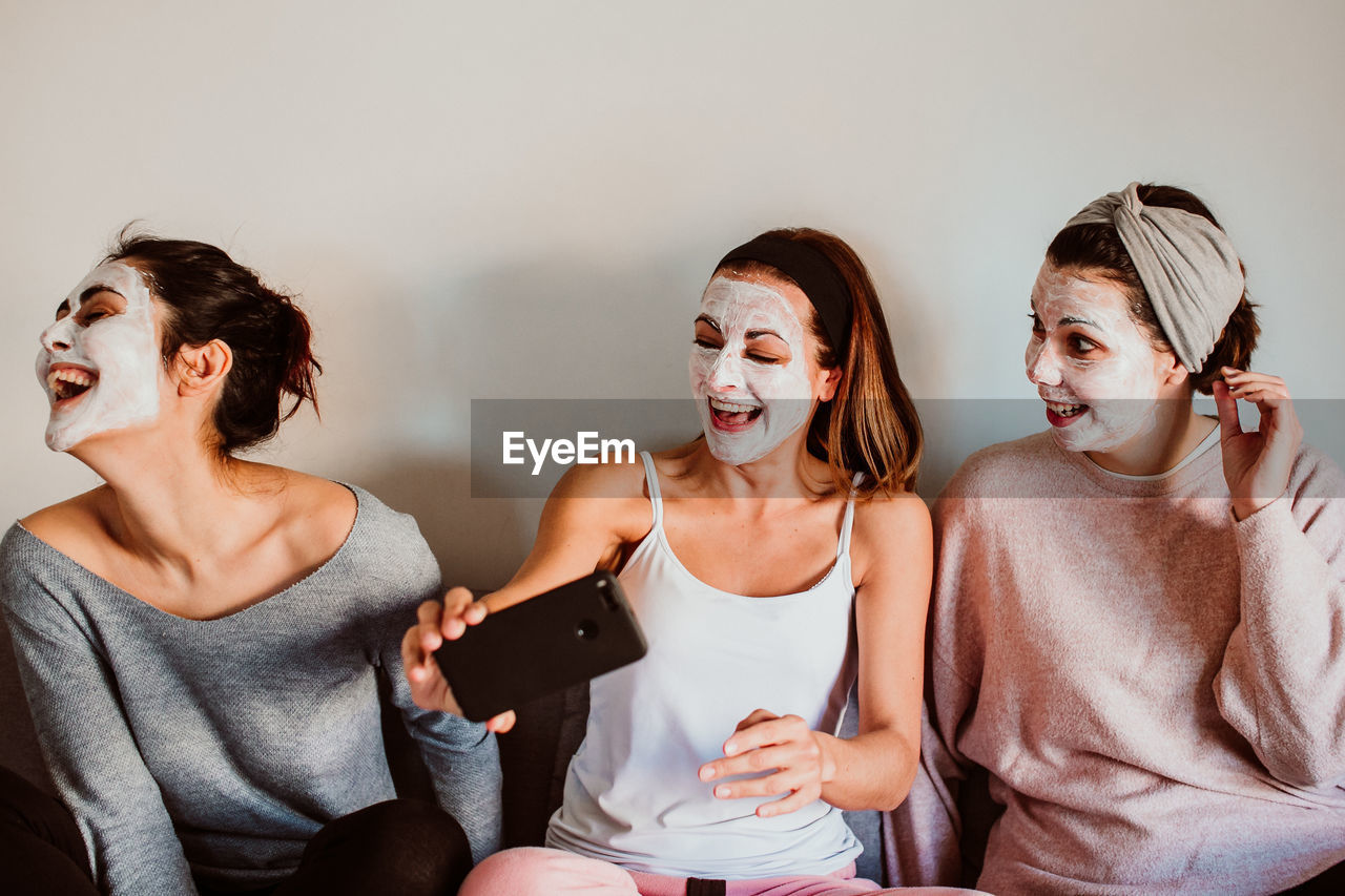 Female friends with facial masks sitting against wall at home