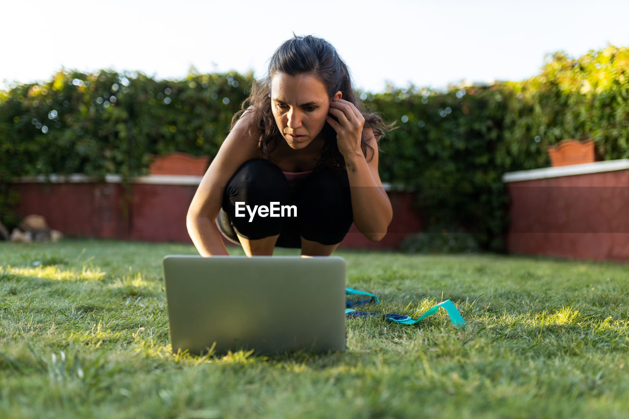 Serious athletic female choosing online tutorial on laptop while preparing for workout in backyard in summer