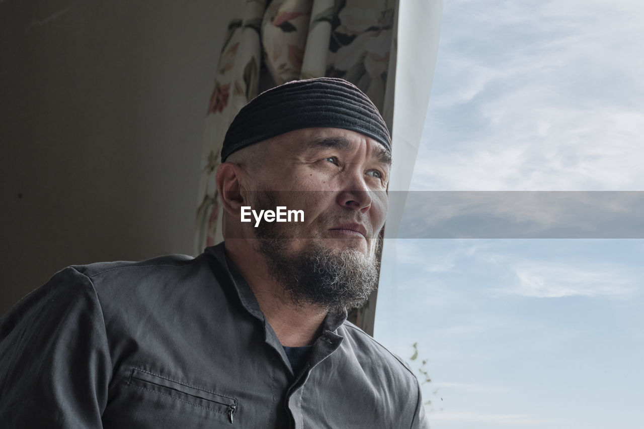 Senior asian muslim man with beard wearing skullcap sitting by window and looking into distance. 