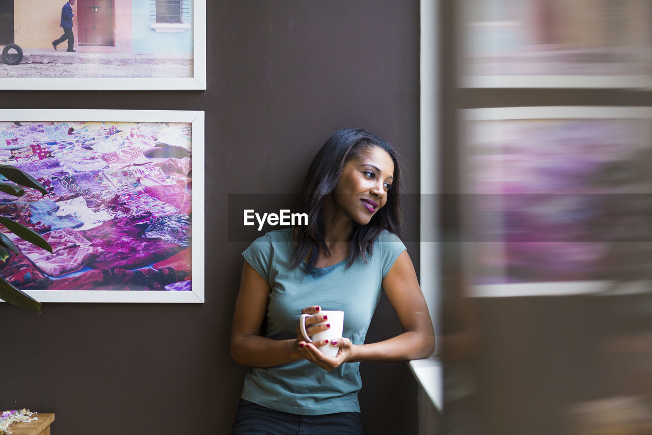 Thoughtful woman holding coffee cup while standing against picture frames on wall at home