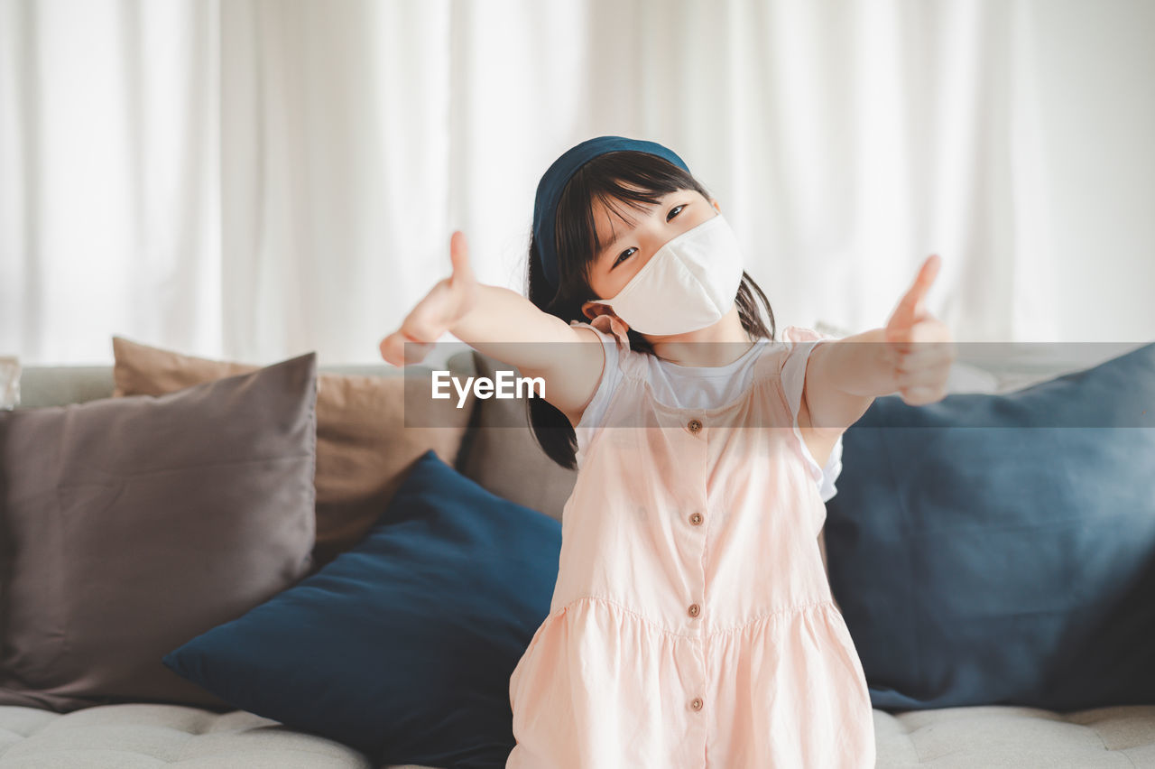 Portrait of girl wearing mask with arms outstretched sitting on sofa