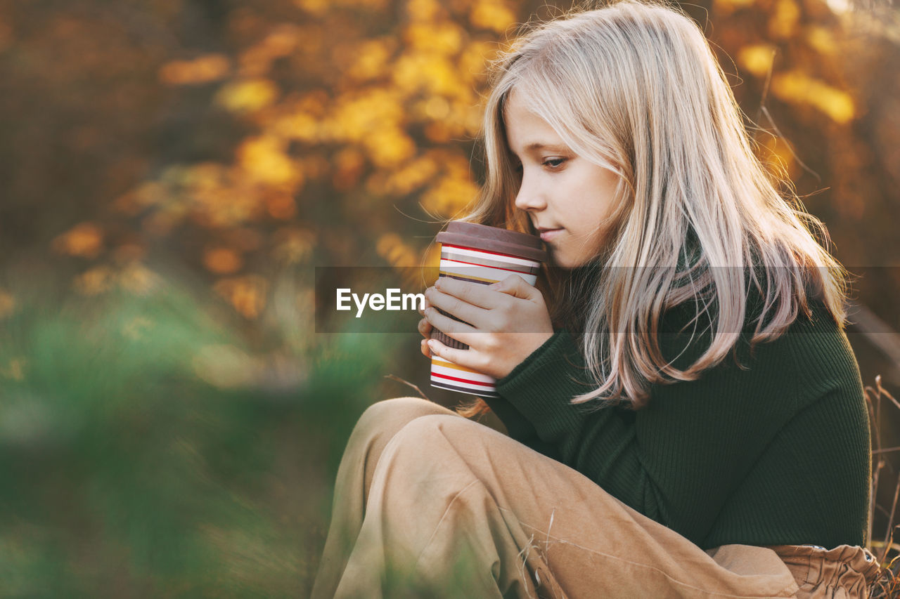 A beautiful teenage girl with blond hair sits thoughtfully in an autumn park, holds a thermos